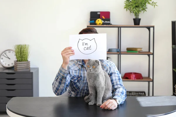 Young man holding paper with drawing and text I'M CAT and his cute pet at home — Stock Photo, Image