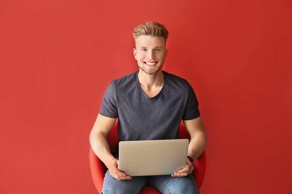 Handsome young man with laptop sitting on chair against color background — Stock Photo, Image