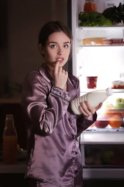 Mujer joven cautelosa con botella de leche cerca del refrigerador por la noche —  Fotos de Stock