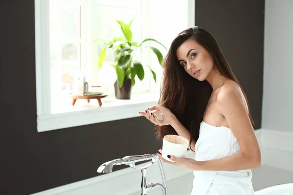 Beautiful young woman applying cosmetics onto her healthy long hair at home — Stock Photo, Image