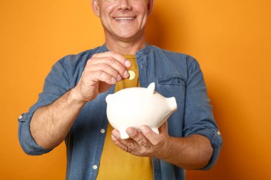 Handsome middle-aged man putting coin into piggy bank on color background clipart