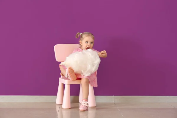 कपास कैंडी के साथ सुंदर छोटी लड़की रंग की दीवार के खिलाफ कुर्सी पर बैठी — स्टॉक फ़ोटो, इमेज