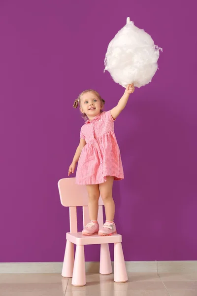 Cute little girl with cotton candy standing on chair against color wall — Stock Photo, Image