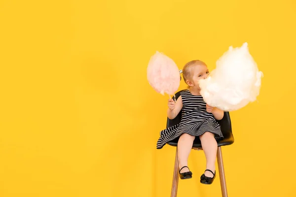 Cute little girl with cotton candy sitting on chair against color background — Stock Photo, Image