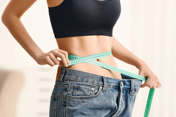 穿着宽松牛仔裤的女人在家里测量她的腰部。减肥概念 — 图库照片
