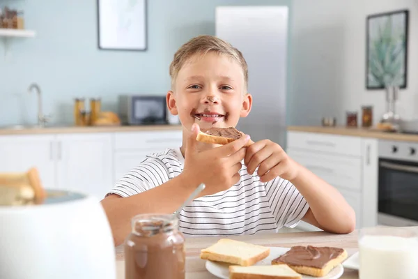 Funny little boy eating tasty toasts with chocolate spreading in kitchen — Stock Photo, Image