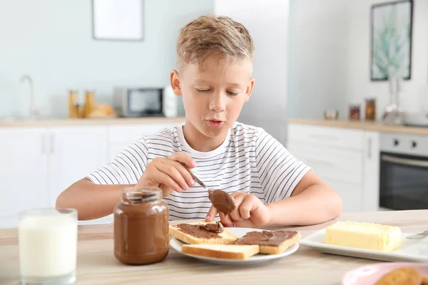 Vicces kisfiú eszik ízletes pirítós csokoládé terjedése a konyhában — Stock Fotó