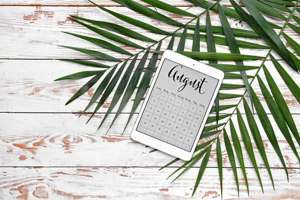 Tablet computer with calendar on screen and tropical leaves on wooden background — Stock Photo, Image
