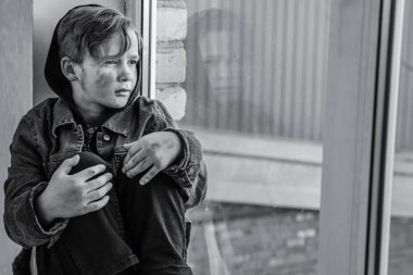 Homeless little boy sitting on window sill indoors clipart