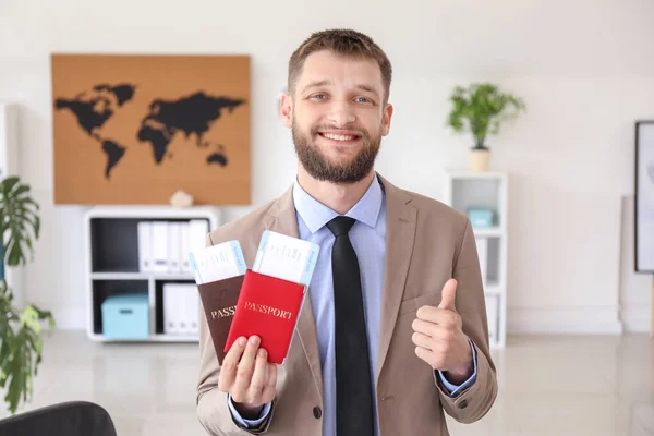 Male travel agent with passports and tickets showing thumb-up gesture in office