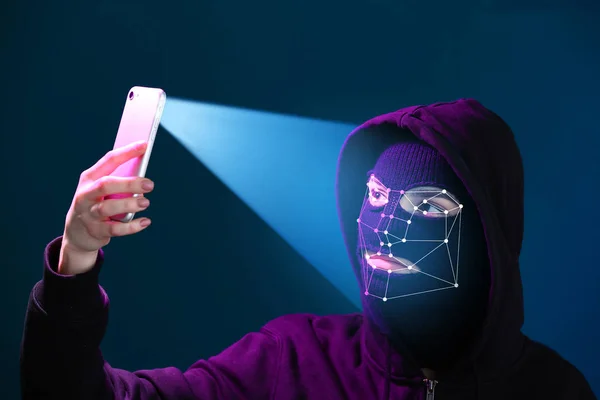 Female hacker in mask holding smartphone protected with facial recognition system