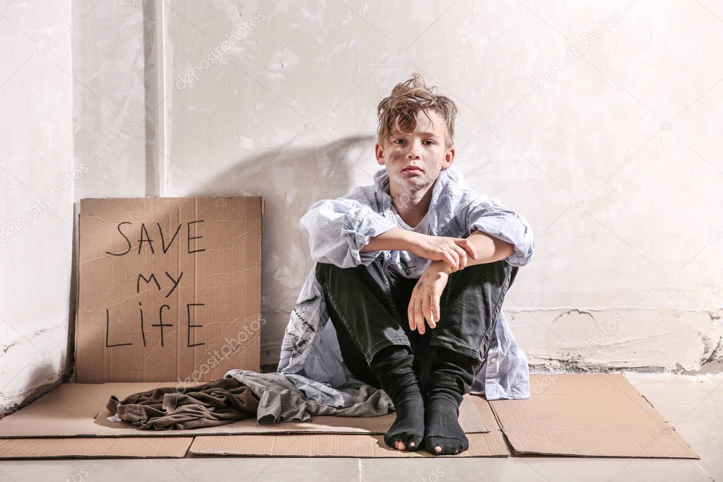 Homeless little boy sitting on floor near wall