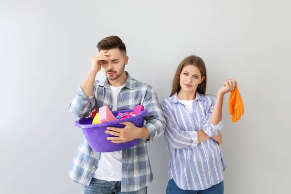 Young wife and stressed husband with cleaning supplies on grey background — Stock Photo, Image