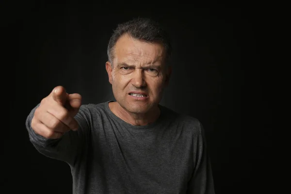 Angry mature man pointing at viewer on dark background — Stock Photo, Image