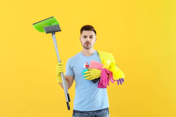 Stressed young man with cleaning supplies on color background