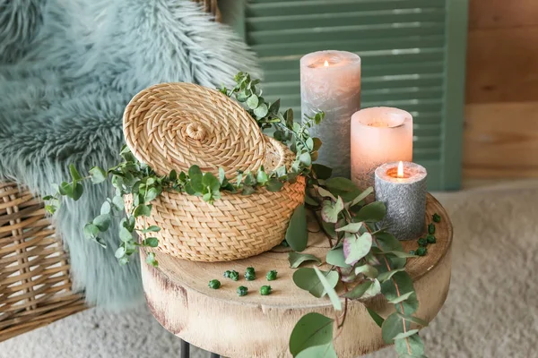 Wicker box, burning candles and eucalyptus branches on table in room