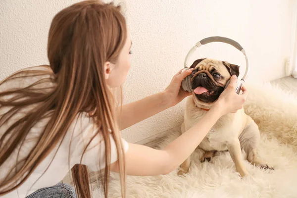 Beautiful young woman putting headphones on cute pug dog at home — Stock Photo, Image
