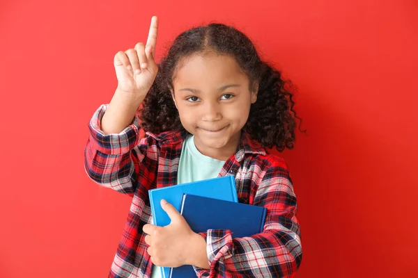 Portrait of adorable little African-American girl with books and raised index finger on color background