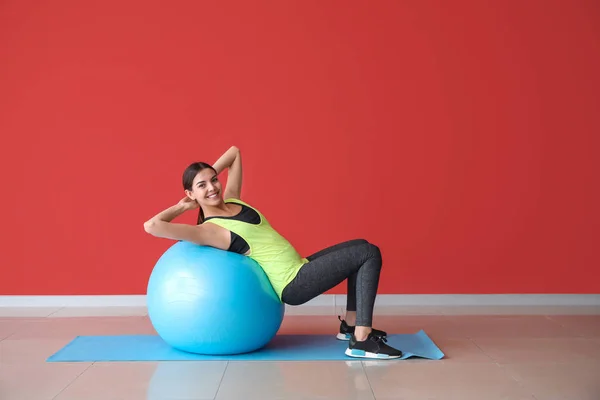 Jovem mulher esportiva com fitball fazendo exercícios perto da parede de cor — Fotografia de Stock