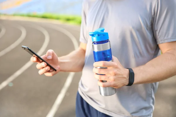 Sporty young man with mobile phone and bottle of water at the stadium — Stock Photo, Image