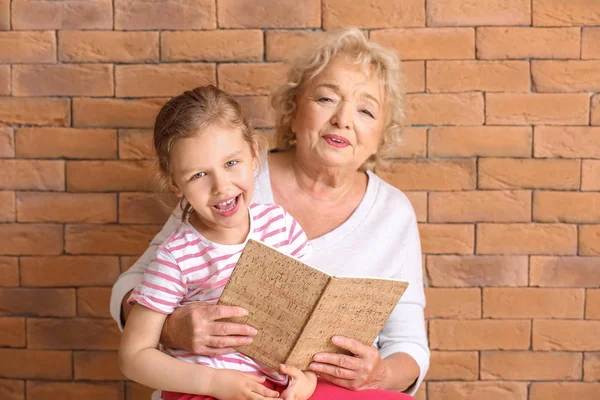 Χαριτωμένο μικρό κορίτσι με τη γιαγιά ανάγνωση βιβλίο εναντίον τούβλο φόντο — Φωτογραφία Αρχείου