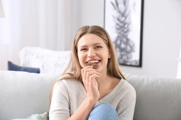 Beautiful young woman with tasty cookie sitting on sofa at home — Stock Photo, Image