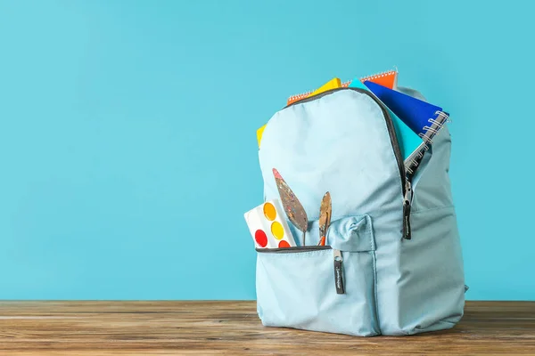 Backpack with school supplies on wooden table against color background — Stock Photo, Image
