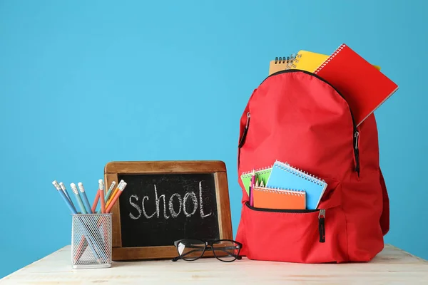 Ryggsäck med förbrukningsmaterial och svarta tavlan med text skola på bordet mot färg bakgrund — Stockfoto