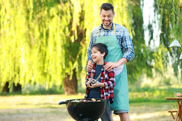 Ragazzino con padre che cucina cibo gustoso sul barbecue all'aperto — Foto Stock