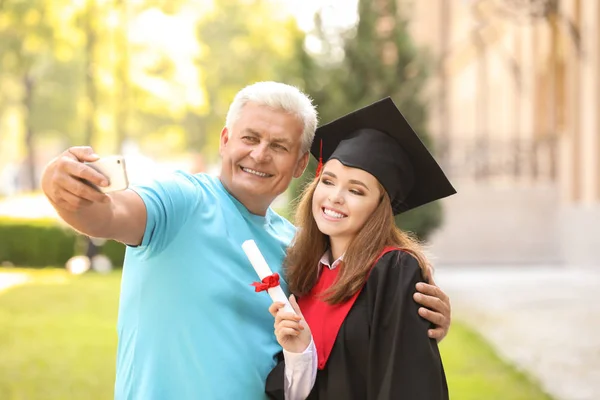 Szczęśliwa młoda kobieta z ojcem biorąc selfie w dniu ukończenia studiów — Zdjęcie stockowe