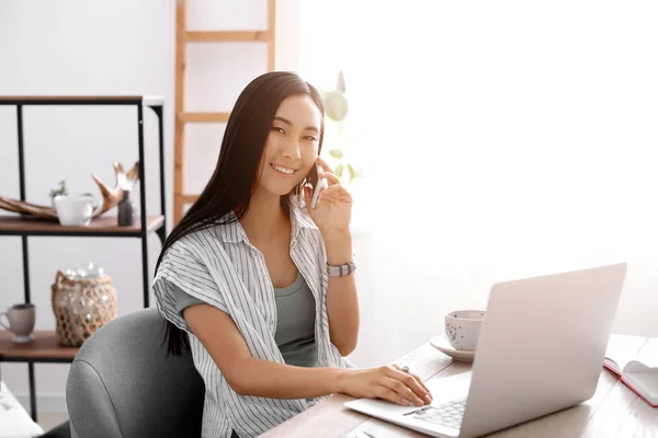 Asian blogger with laptop talking by phone at table — Stock Photo, Image