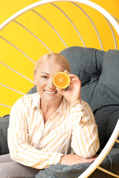 Beautiful mature woman with half of orange fruit sitting in armchair on color background — Stock Photo, Image