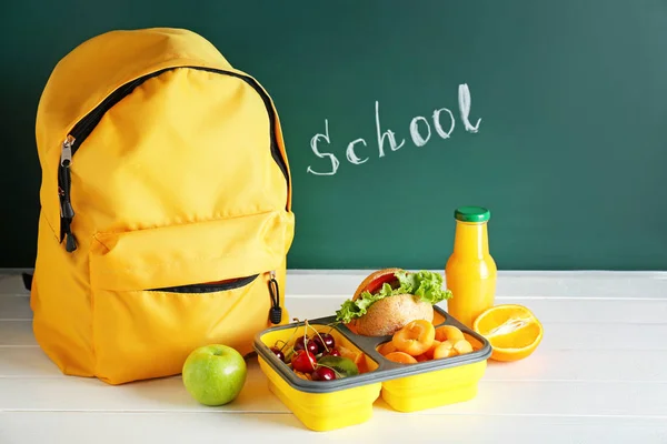 School lunch box with tasty food and backpack on table in classroom