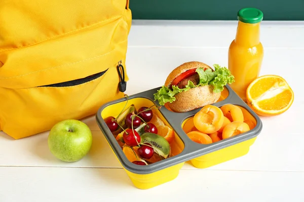 Pudełko na lunch w szkole ze smacznym jedzeniem i plecakiem na stole w klasie — Zdjęcie stockowe