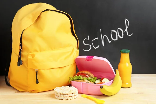 School lunch box with tasty food and backpack on table in classroom — Stock Photo, Image