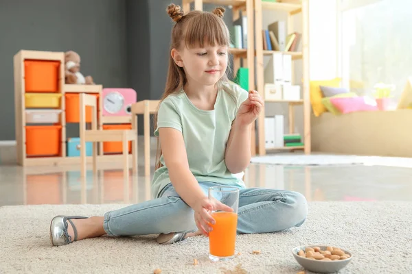 Careless little girl eating nuts and drinking juice while sitting on carpet — Stock Photo, Image