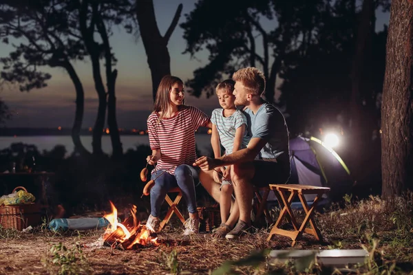 Family roasting sausages over campfire in evening