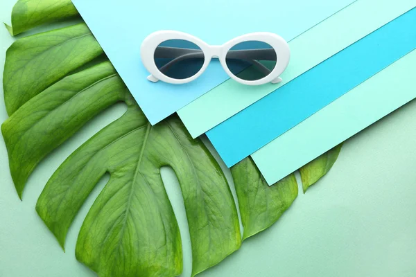 Stylish sunglasses with papers and tropical leaf on color background — Stock Photo, Image