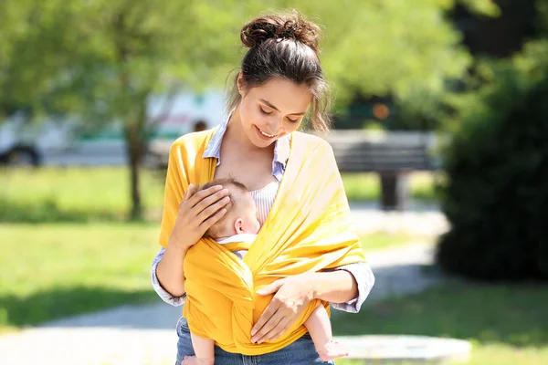 Young mother with her baby in sling walking in park — Stock Photo, Image