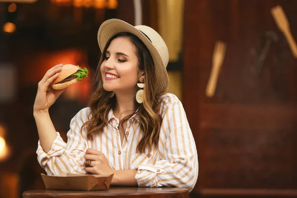 Красивая молодая женщина ест вкусный бургер в кафе на открытом воздухе — стоковое фото