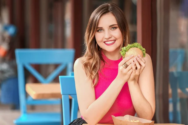 Красивая молодая женщина ест вкусный бургер в кафе на открытом воздухе — стоковое фото