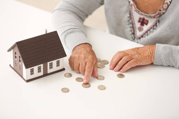 Elderly woman with figure of house counting coins on table, closeup. Concept of payment for utility services