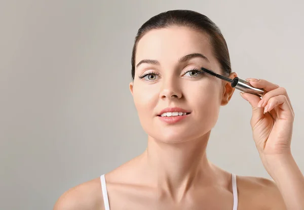 Beautiful young woman with eyelash extensions applying mascara against grey background — Stock Photo, Image