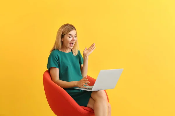 Schockierte Frau mit Laptop sitzt im Sessel auf farbigem Hintergrund — Stockfoto