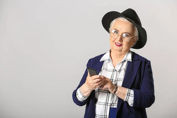 Stylish elderly woman with mobile phone on grey background