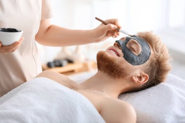 Kozmetik uzmanı spa salonunda adamın yüzüne maske takıyor.