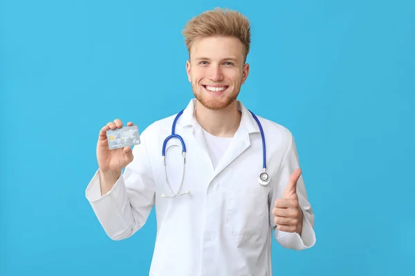 Médico do sexo masculino feliz com cartão de crédito mostrando gesto de polegar para cima no fundo de cor — Fotografia de Stock