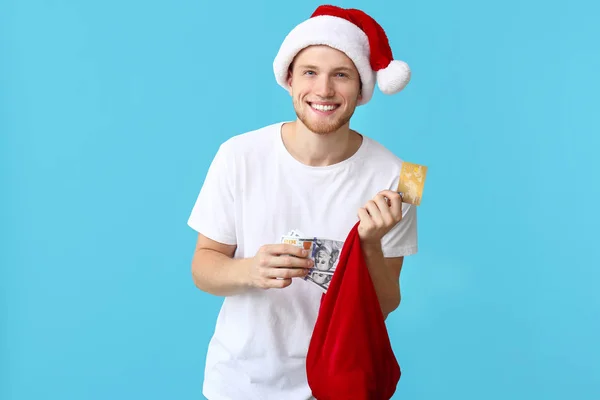 Happy young man in Santa hat, with credit card, money and bag on color background — Stock Photo, Image