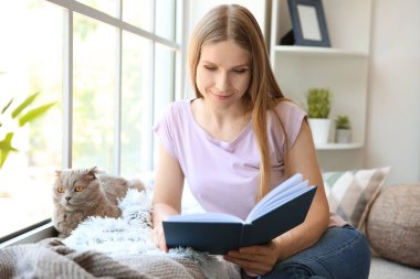 Evde pencere yakınında sevimli kedi okuma kitabı ile güzel kadın