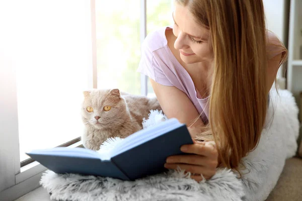 Όμορφη γυναίκα με χαριτωμένο βιβλίο ανάγνωσης γάτα κοντά στο παράθυρο στο σπίτι — Φωτογραφία Αρχείου